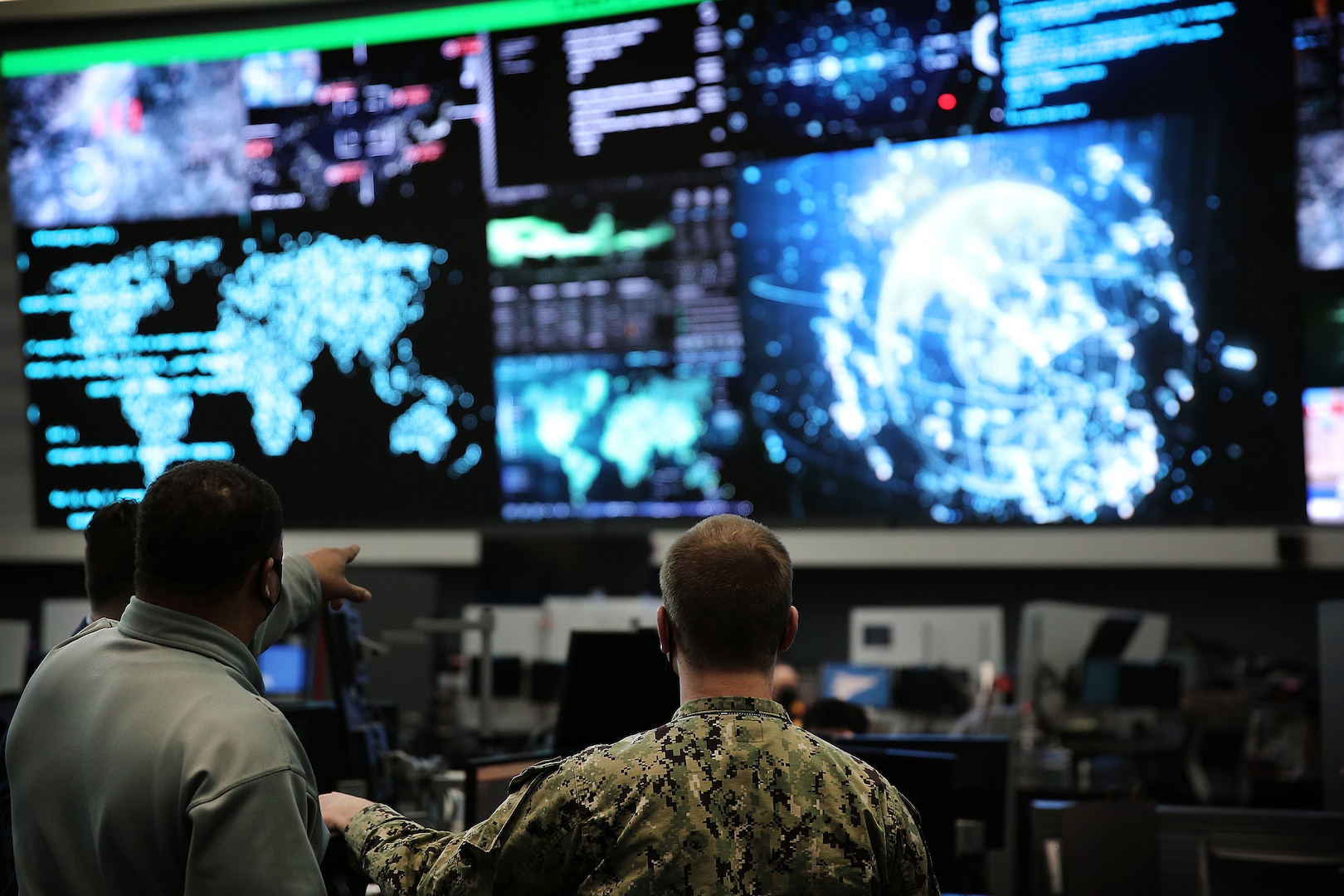 Two men standing in a security operations center. One is pointing at a screen, the other wearing a uniform observing.